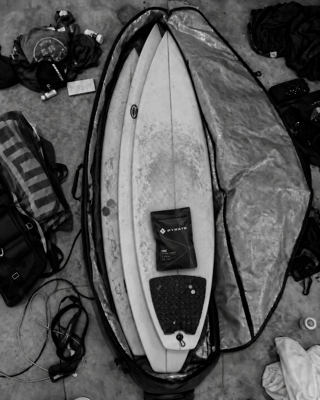PYRATE Edge pouch resting on a surfboard inside an open travel bag with packed surf gear.
